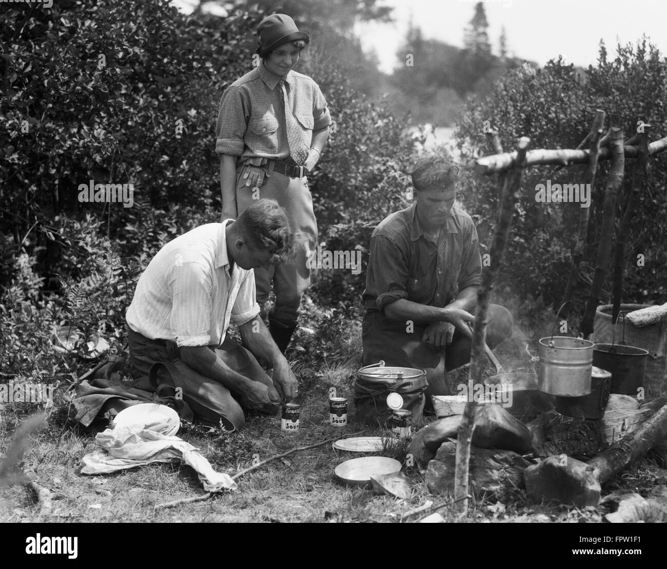 1920s WOMAN WATCHING TWO MEN AROUND CAMPFIRE ONE COOKING IN SKILLET OTHER OPENING CANS OF SOUP MERSEY RIVER NOVA SCOTIA CANADA Stock Photo