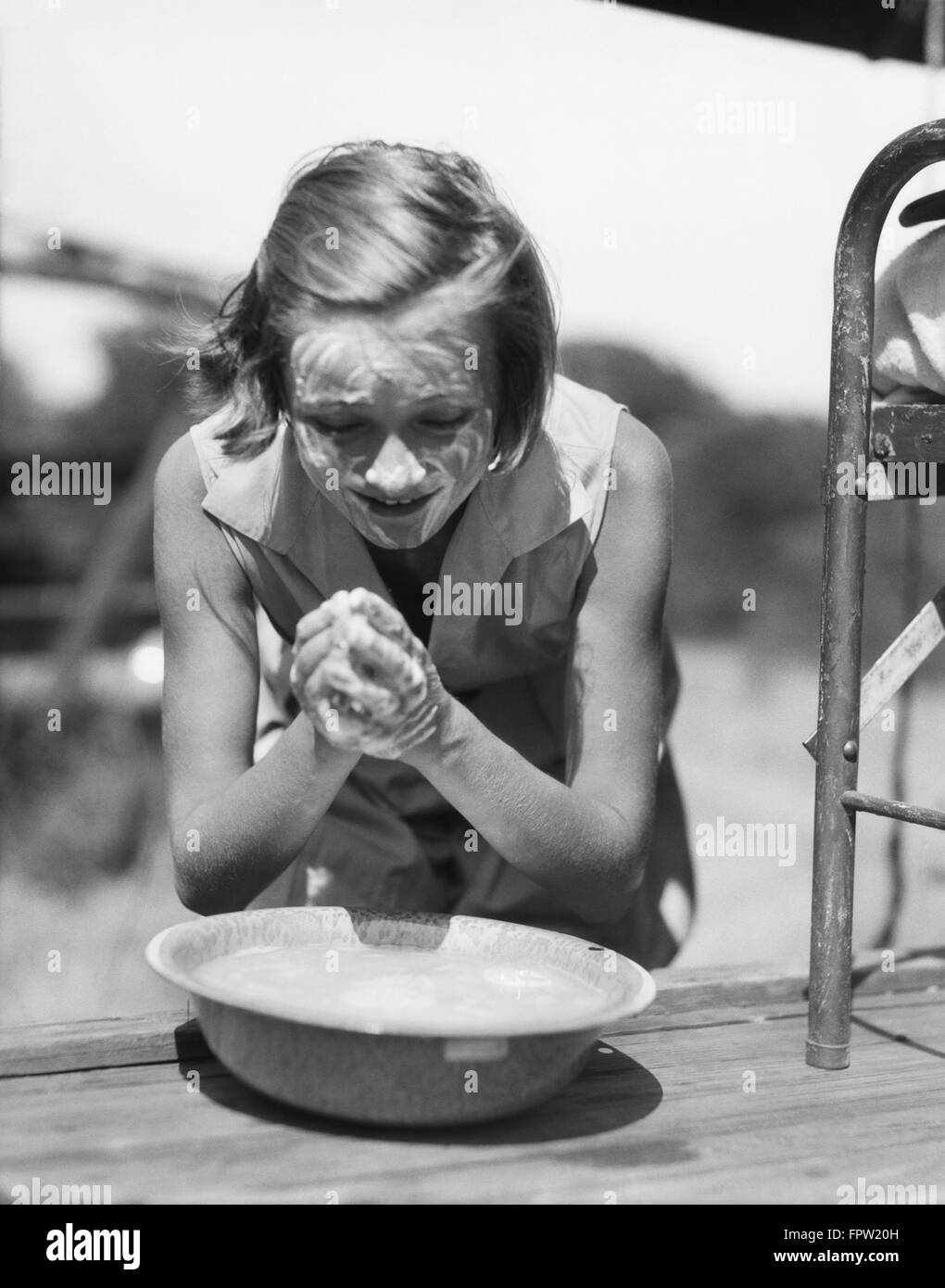 1930s YOUNG GIRL LEANING OVER BOWL WASHING HANDS AND FACE WITH SOAP AT SUMMER CAMP OUTDOORS Stock Photo