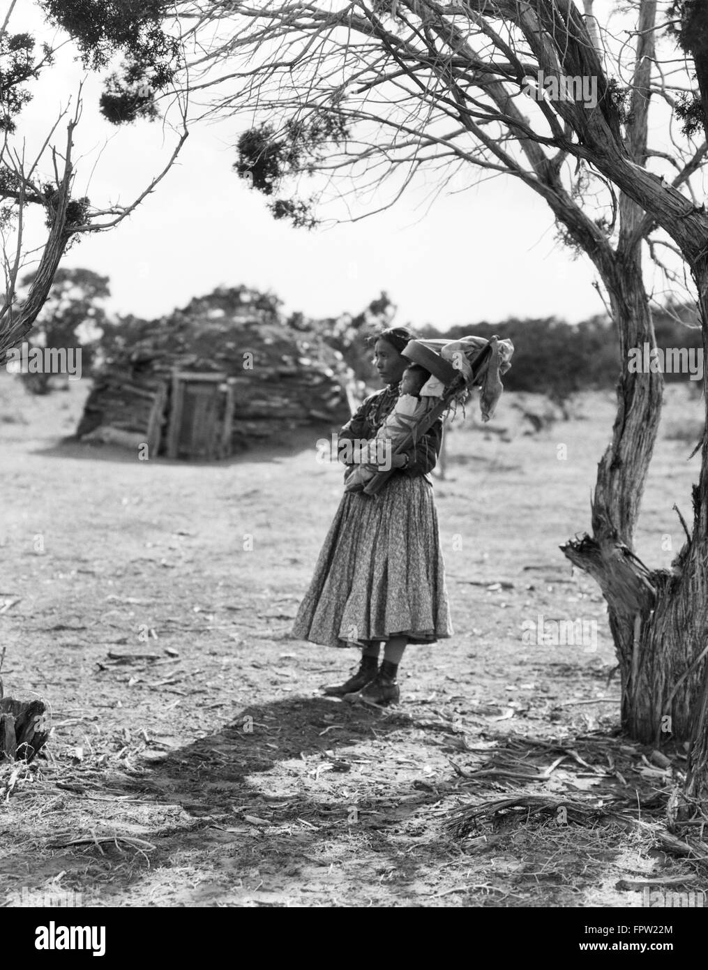 1930s NATIVE AMERICAN NAVAJO WOMAN HOLDING BABY PAPOOSE UNDER TREES WITH HOGAN IN BACKGROUND Stock Photo