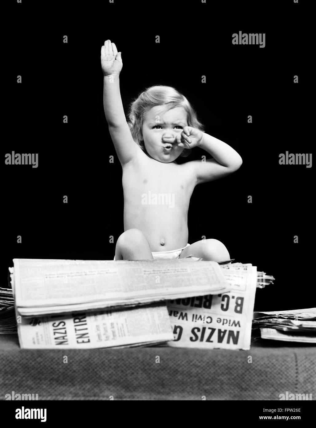 1940s LITTLE BLOND TODDLER GIRL IMITATING HITLER GIVING NAZI SALUTE SITTING ON PILE OF NEWSPAPERS Stock Photo