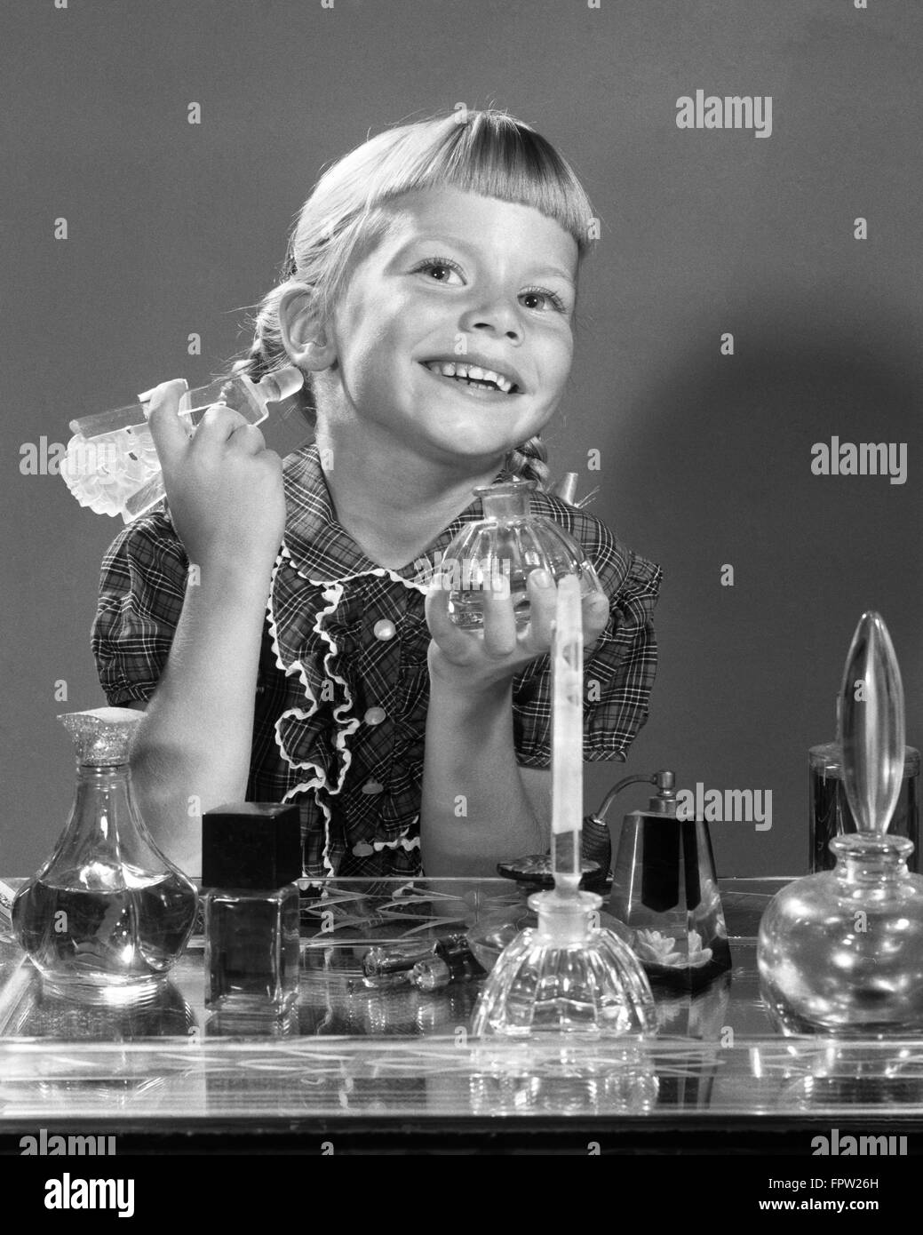 1960s SMILING LITTLE GIRL SITTING AT MOTHER’S BUREAU VANITY PERFUME BOTTLES IN FRONT LOOKING AT CAMERA IN MIRROR Stock Photo