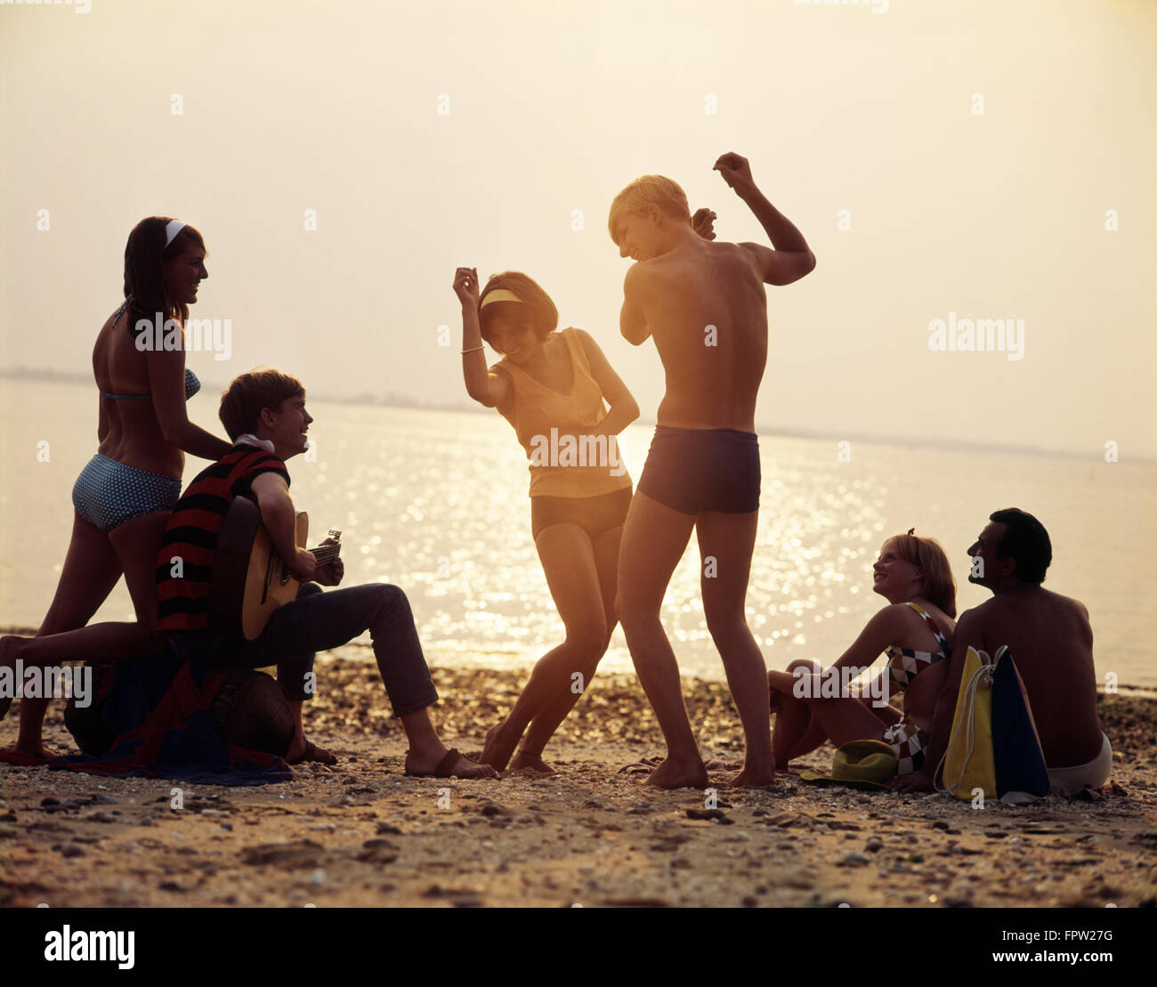 1960s GROUP OF YOUNG COUPLES PLAYING GUITAR AND DANCING SUNSET BEACH PARTY Stock Photo