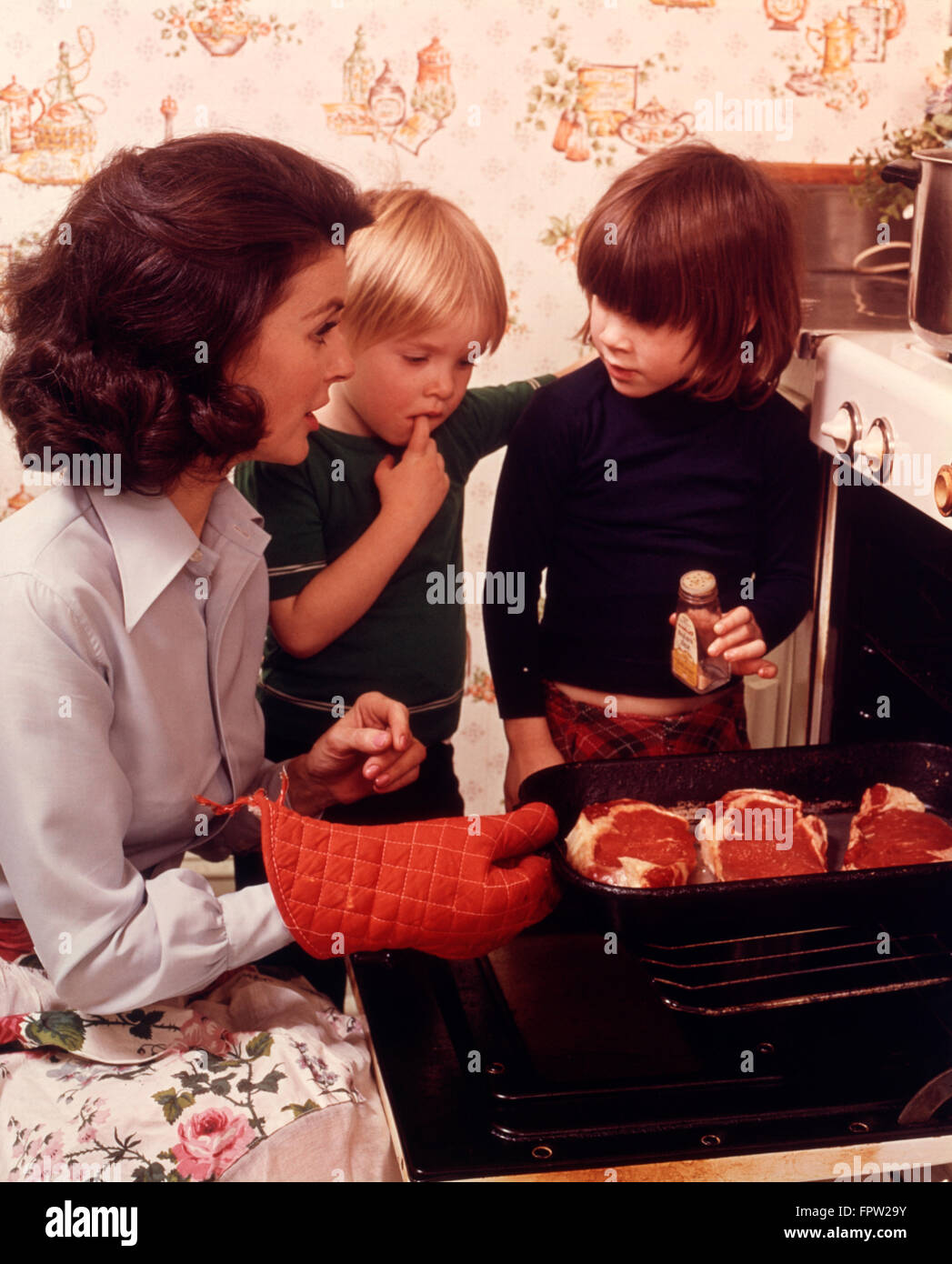 1970s WOMAN MOTHER 2 CHILDREN BOY GIRL KITCHEN OVEN DOOR OPEN CHECKING STEAKS MEAT ADDING SPICE COOKING Stock Photo