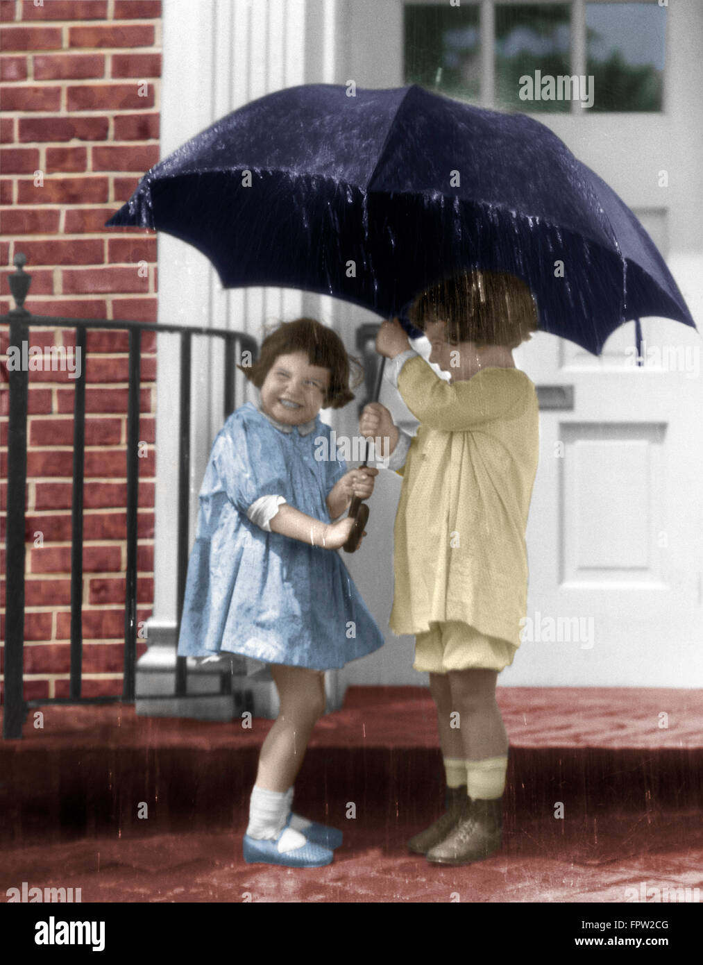 1920s PAIR OF LITTLE GIRLS STANDING ON FRONT STOOP UNDER UMBRELLA IN POURING RAIN Stock Photo