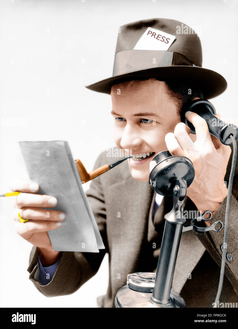 1930s EXCITED MAN NEWSPAPER REPORTER SMOKING PIPE PRESS PASS IN HAT TALKING ON UPRIGHT PHONE REPORTING STORY FROM NOTE PAD Stock Photo