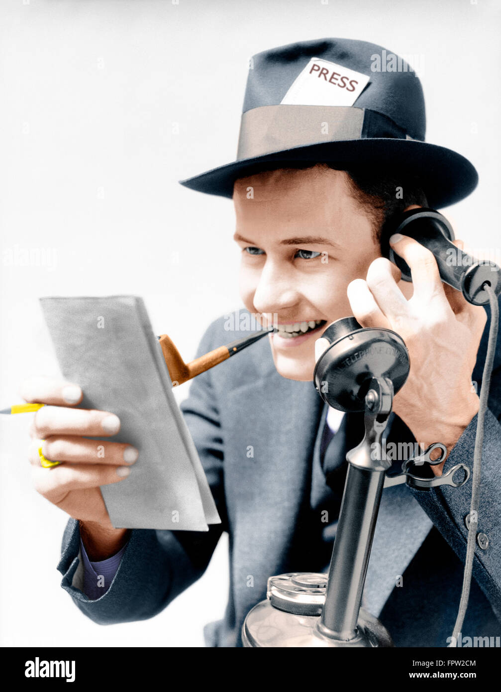 1930s EXCITED MAN NEWSPAPER REPORTER SMOKING PIPE PRESS PASS IN HAT TALKING ON UPRIGHT PHONE REPORTING STORY FROM NOTE PAD Stock Photo