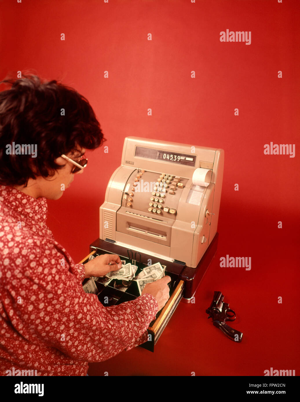 1970s MAN ROBBER ARMED WITH GUN WEARING SUNGLASSES BLACK WIG AND RED PRINT SHIRT STEALING MONEY FROM CASH REGISTER DRAWER Stock Photo