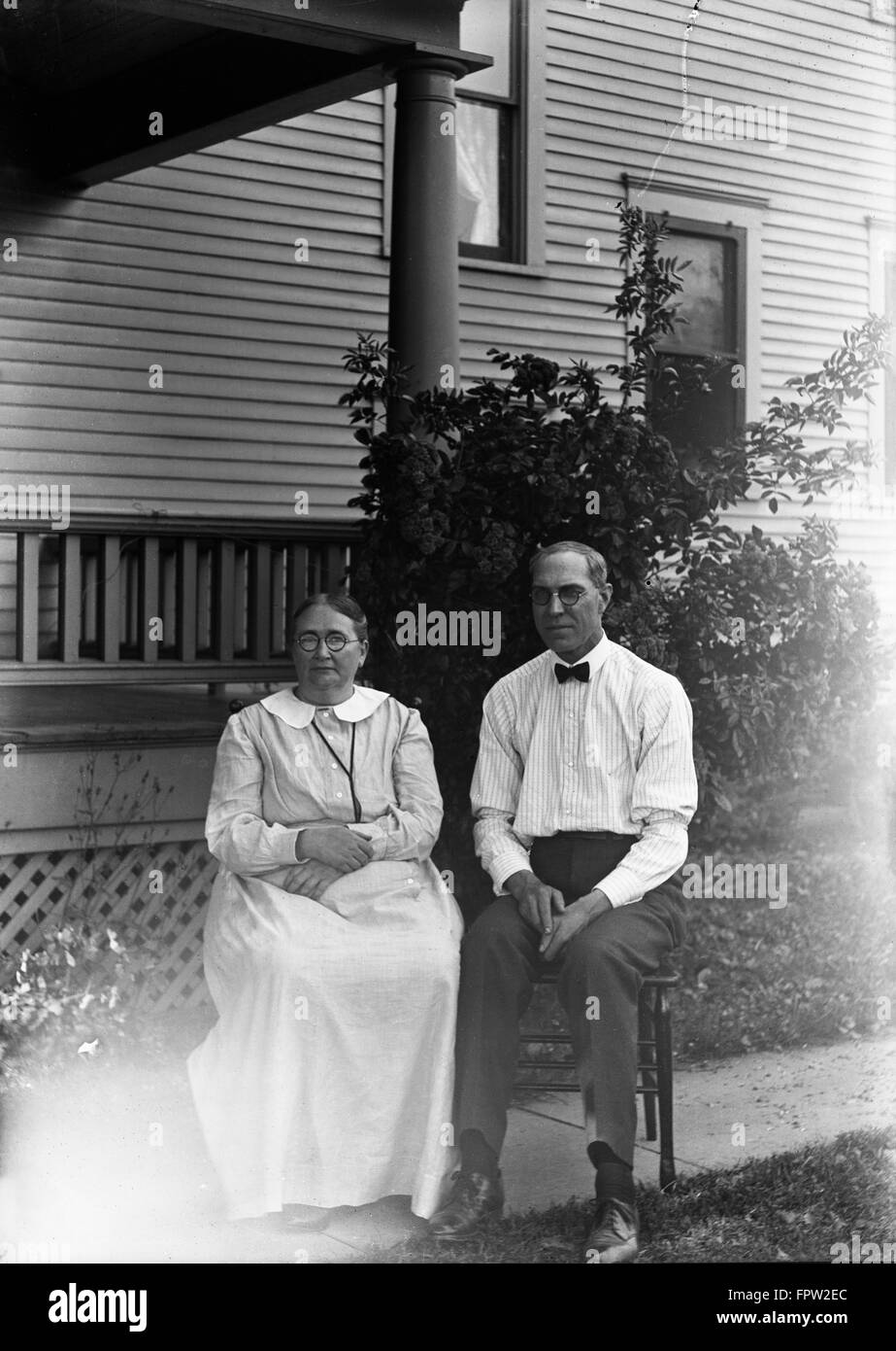 1900s PORTRAIT COUPLE MAN WOMAN SITTING ON CHAIRS IN FRONT OF HOUSE LOOKING AT CAMERA Stock Photo