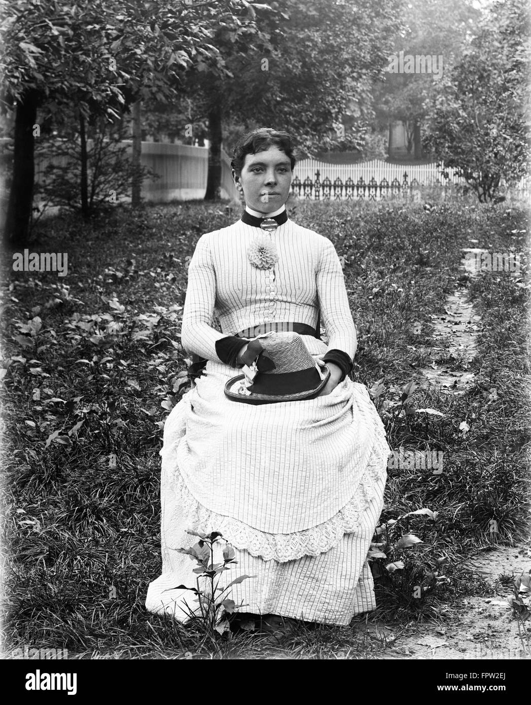 1890s 1900s PORTRAIT WOMAN WEARING STRIPED DRESS SITTING IN CHAIR IN GARDEN HOLDING HAT IN LAP Stock Photo