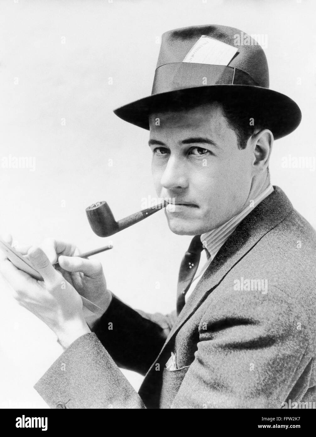 1930s 1940s MAN REPORTER WEARING HAT WITH PRESS PASS WRITING NOTES IN PAD TABLET SMOKING PIPE LOOKING AT CAMERA Stock Photo
