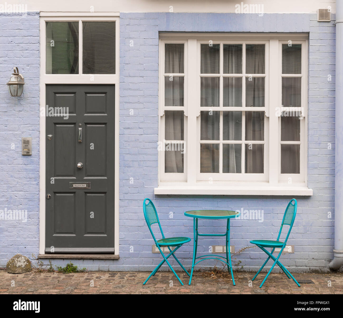 Two blue metal chairs and table standing outside the entrance of an house with black door, white window and lilac brick wall Stock Photo