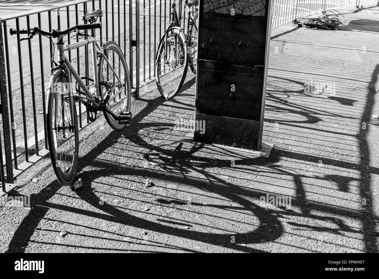 Two road city bikes locked to a metal fence. next to a road. Big shadows projected on the pavement.Black and white. Stock Photo