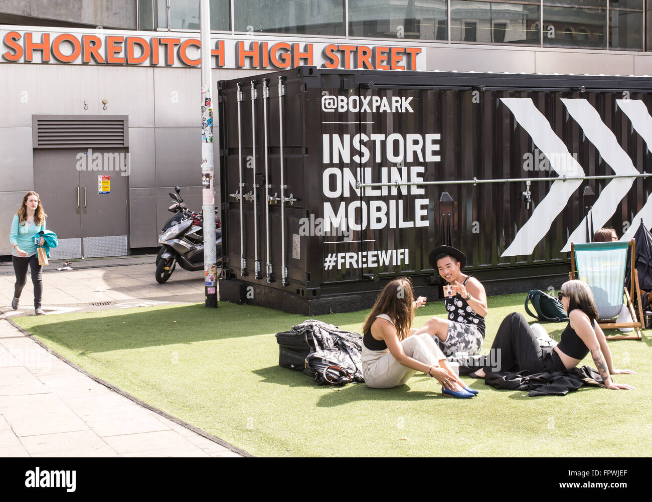 Group of young hipsters sitting down on a fake grass carpet in the Boxpark, the world's first pop-Up mall based in Shoreditch. Stock Photo