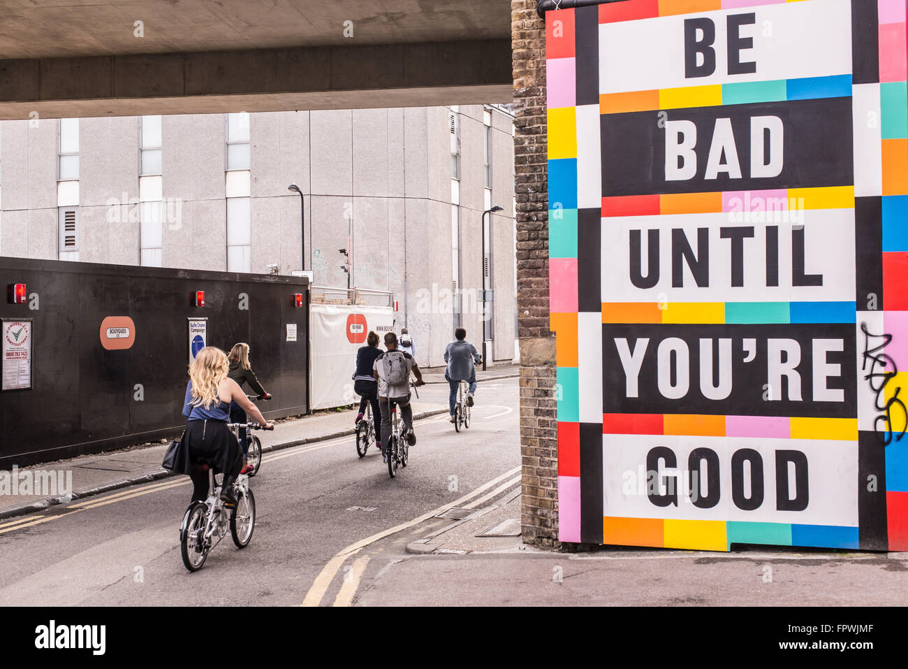 Group of urban cyclists riding next to a big colored billboard with the words 'Be bad until you're good' on it. Stock Photo