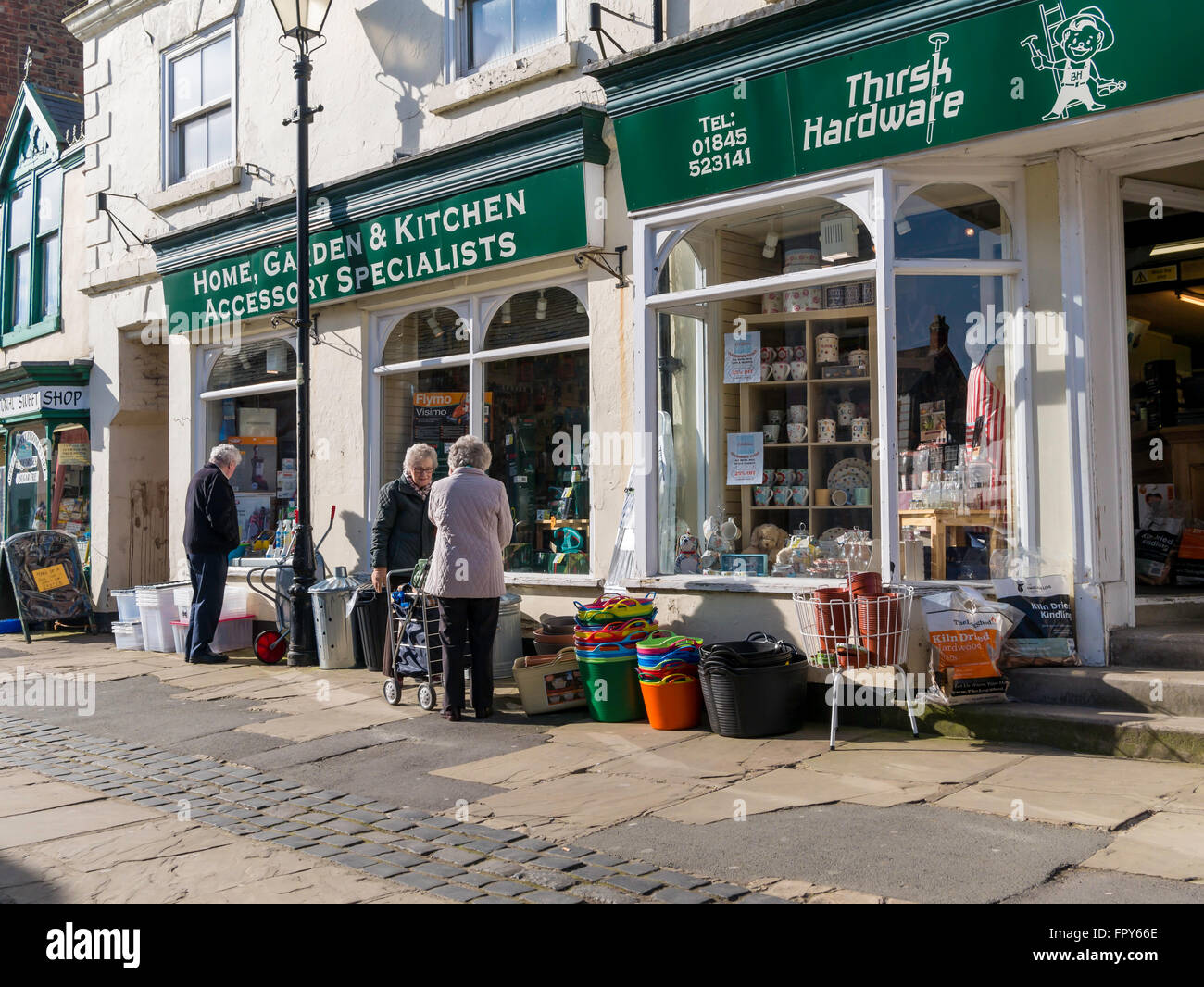 Women talking outside outside a hardware shop with goods displayed in a pedestrianised area of Thirsk Market Place Yorkshire Stock Photo