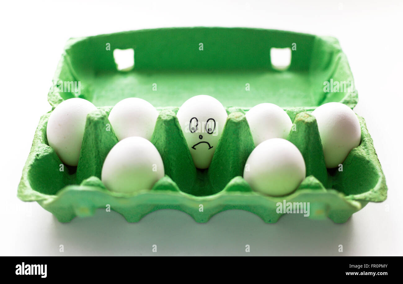 Sad face hand drawn on a white egg. The egg sits in a green cardboard carton surrounded by other faceless eggs. Copy space area Stock Photo