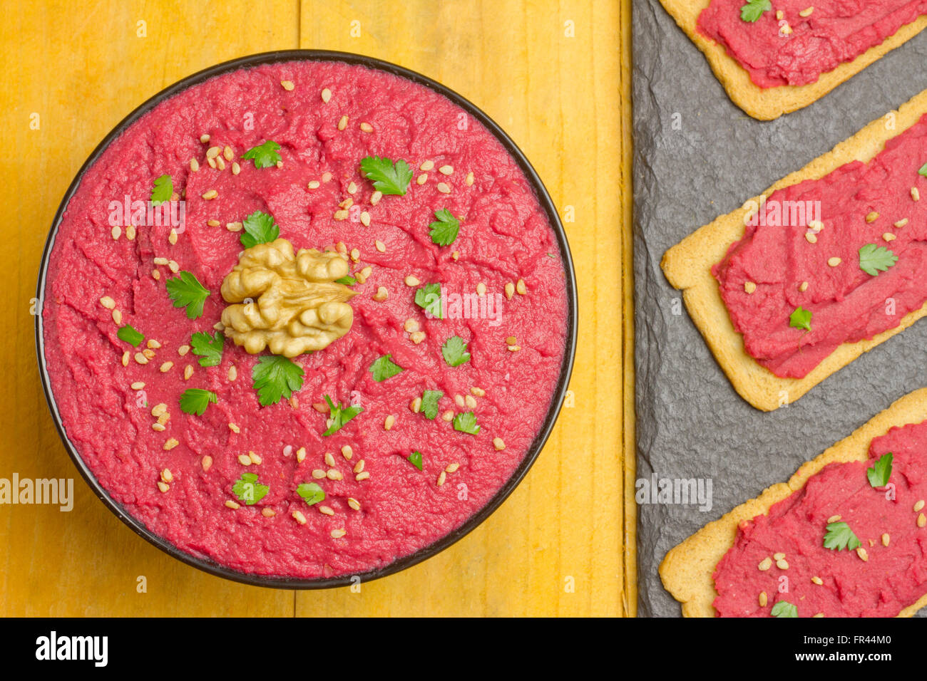 Beet hummus in a black bowl and three slices of bread with beet hummus on a yellow table Stock Photo