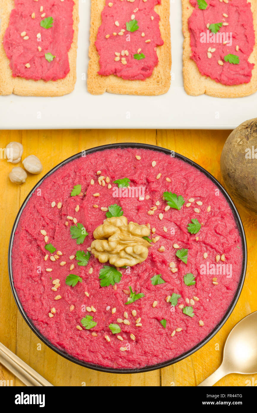Beet hummus in a black bowl, bread slices with beet humus, chickpeas and a fresh beet on a yellow table Stock Photo
