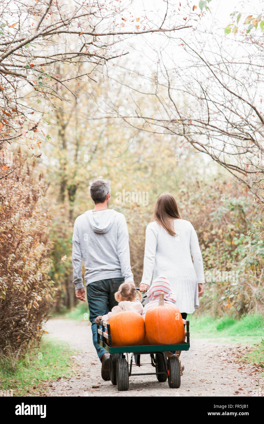 Parents pulling toddler children and pumpkins on wagon in park Stock Photo