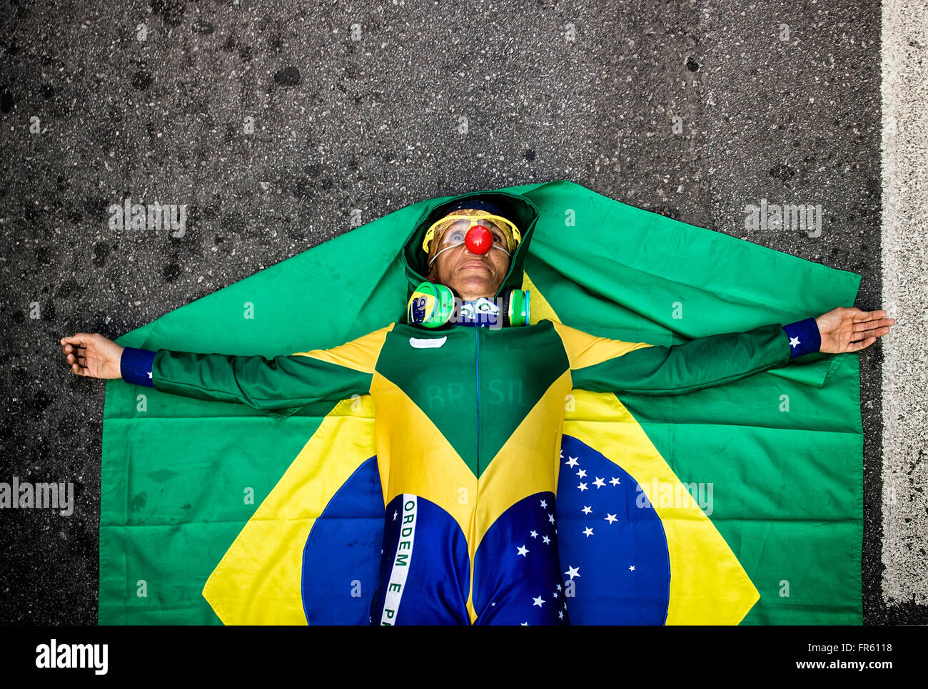 Sao Paulo, Brazil. 20 March 2016. Brazilian woman Ana Luiza Garcez is laying on the ground atop a Brazilian flag with her arms outstretched. She's wearing a Brazilian flag jumpsuit and a clown nose. All over Brazil, people have been taking to the streets to protest the growing number of corruption scandals being uncovered at all levels of the current government. Stock Photo