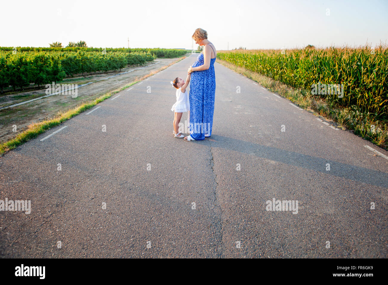 Pregnant mother and her little daughter expecting newborn brother. Country road between cornfields Stock Photo