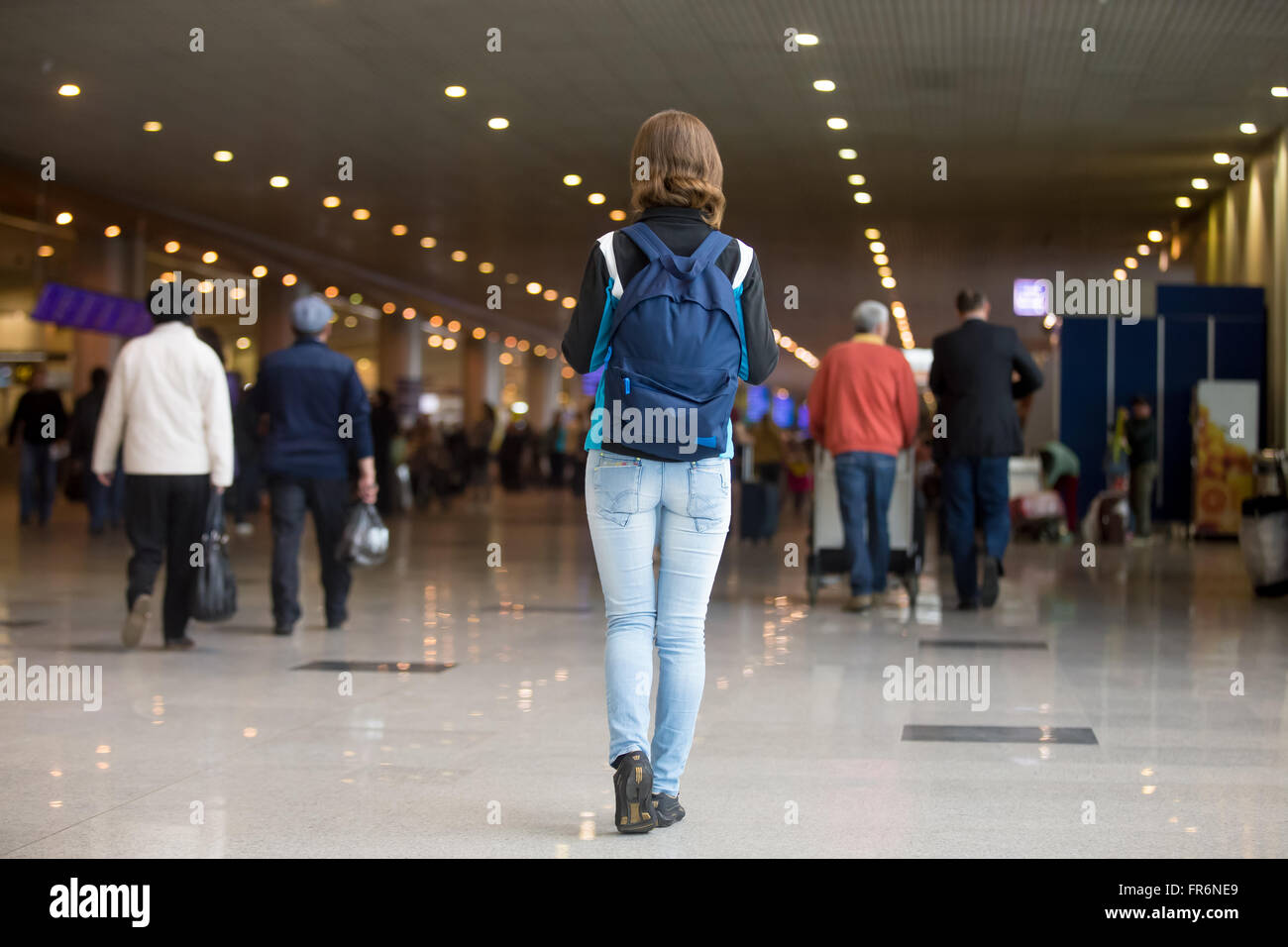 Young woman in 20s with backpack walking in airport terminal, wearing jersey, jeans and sneakers, blurred crowd of travelling pe Stock Photo