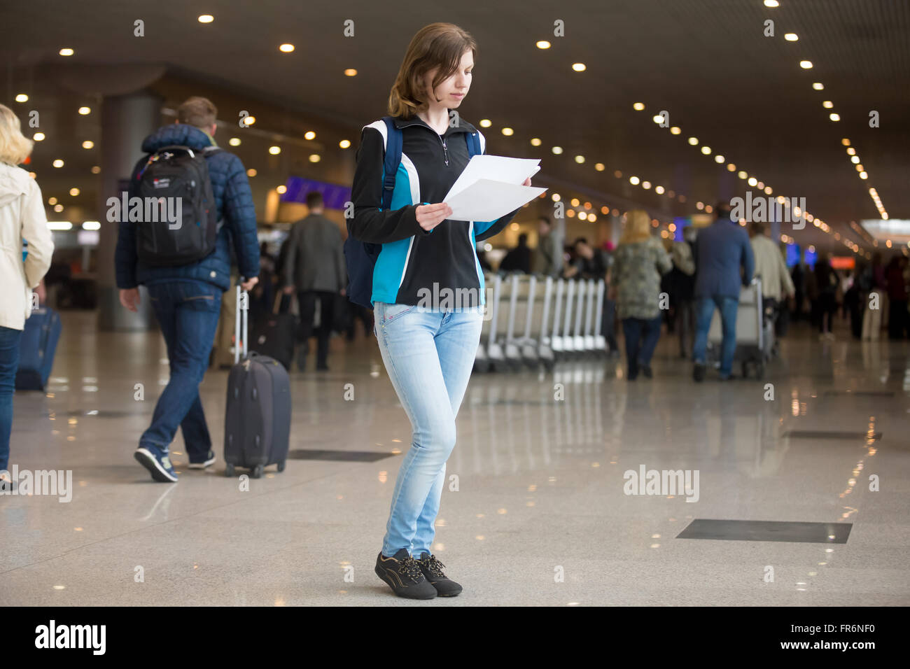 Portrait of young woman with backpack standing in airport terminal, with printed electronic tickets, using cell phone to check i Stock Photo