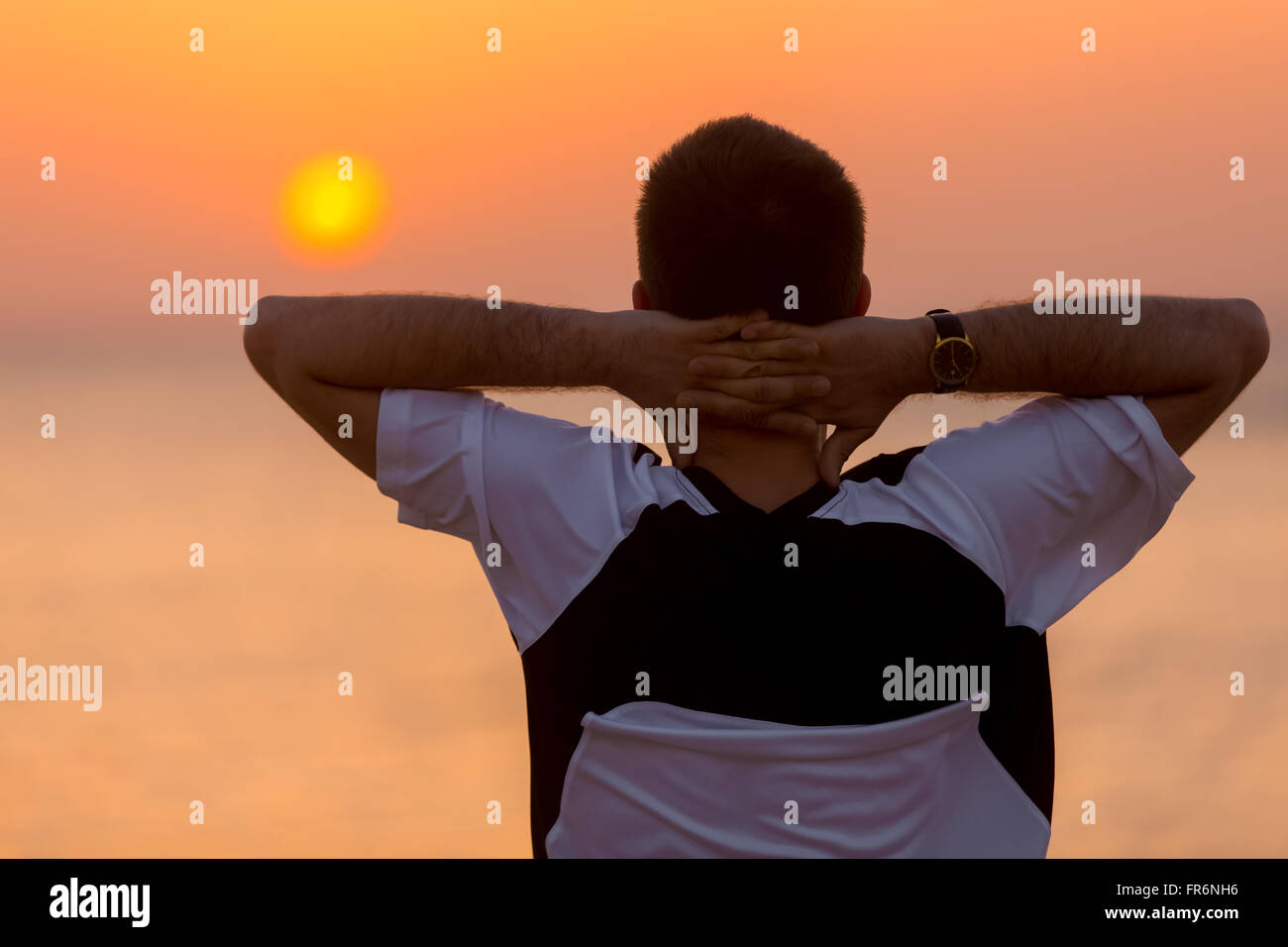 Young man standing at the seashore against colorful sunrise or sunset sky in carefree pose with hands behind his head, enjoying Stock Photo