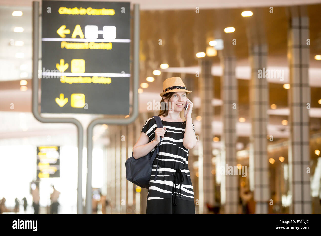Happy smiling young woman in straw summer hat waiting for flight in modern airport terminal building, holding mobile phone, maki Stock Photo