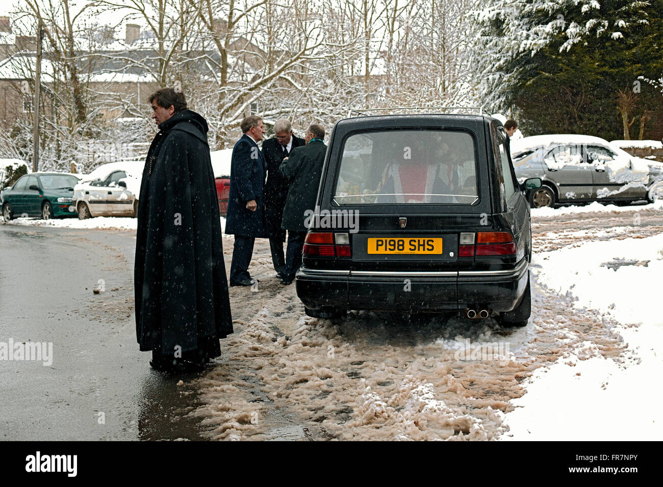 A priest in a long black cloak waits in snow by a hearse following a funeral in Wales Stock Photo