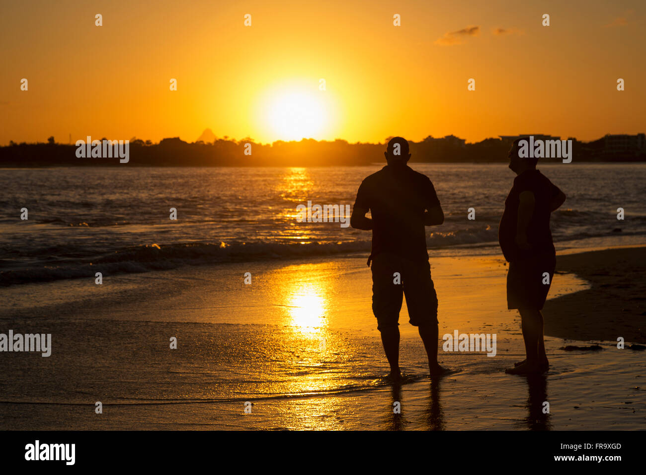 Two men stand in the tide water on a beach at sunset; Caloundra, Queensland, Australia Stock Photo