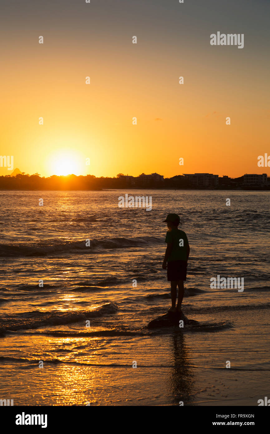 Silhouette of a young boy standing at the water's edge at sunset; Caloundra, Queensland, Australia Stock Photo