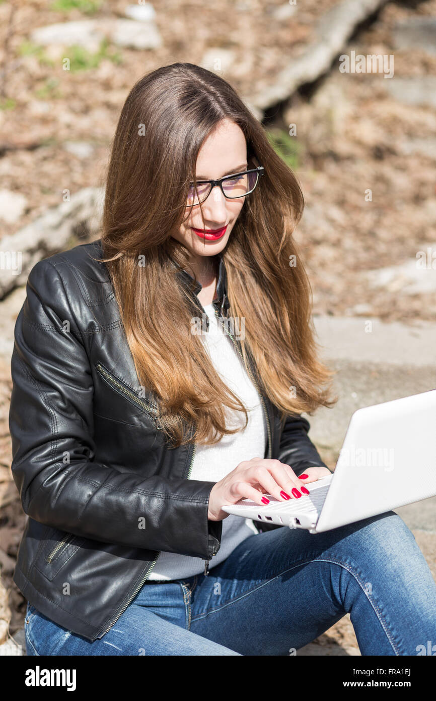 Young woman in glasses and leather jacket using laptop on the stairs. Pretty caucasian girl sitting on stairs working on noteboo Stock Photo
