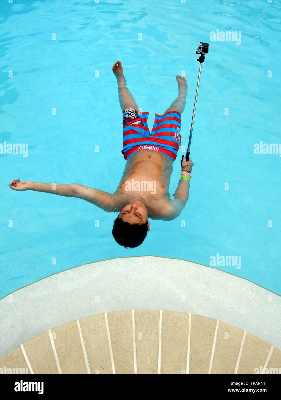 Colorful, graphic image of 12 year old boy lying on his back in a swimming pool, holding a go pro camera on a camera stick . Stock Photo