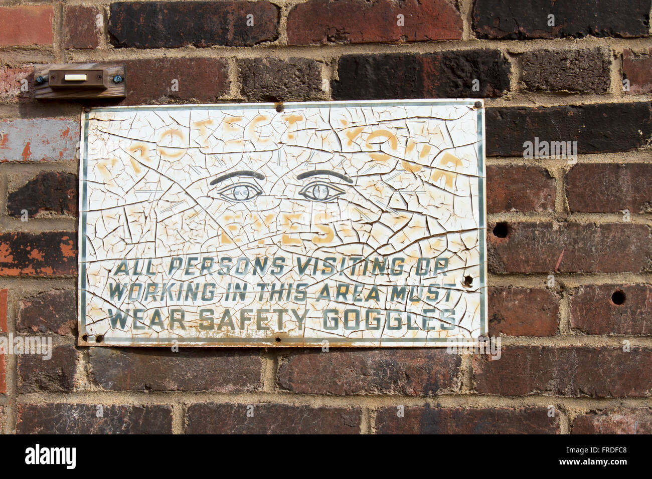 Worn and weathered vintage safety goggles sign on wall of old brick building. Stock Photo