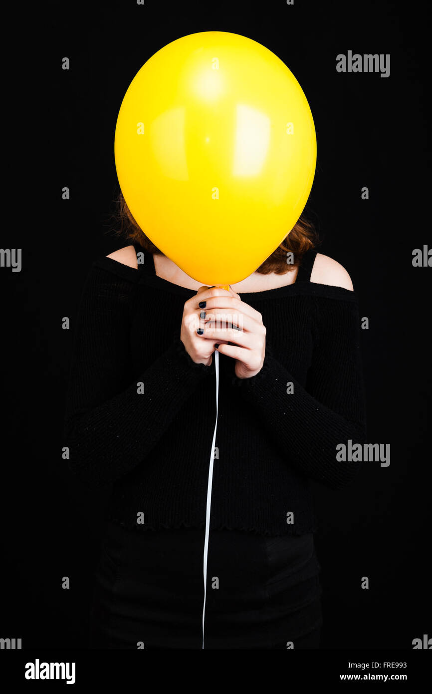 Girl hiding her face under yellow balloon, vertical studio shot over black background Stock Photo