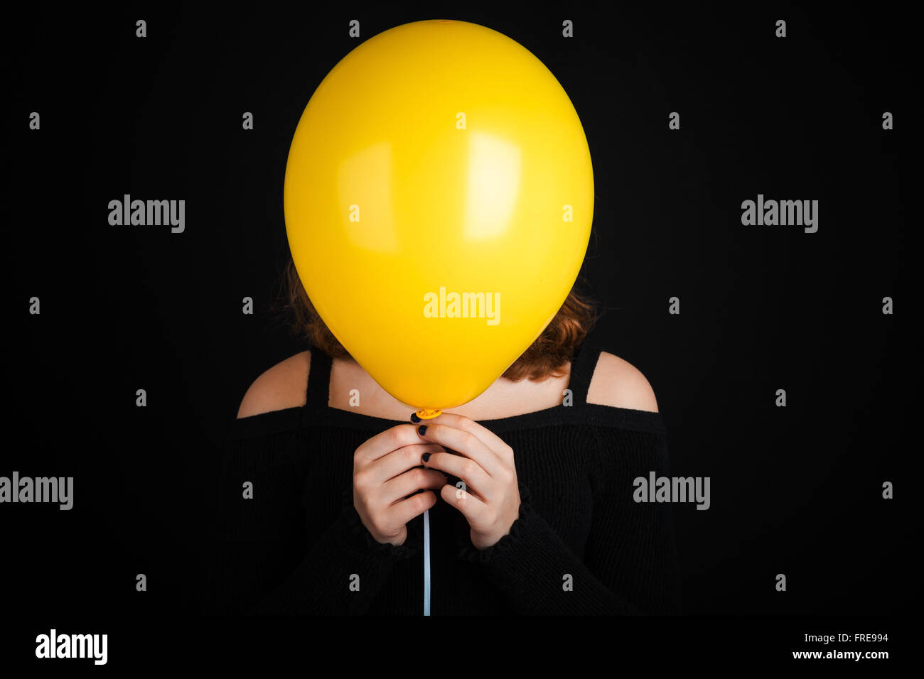Girl hiding her face under yellow balloon, studio shot over black background Stock Photo