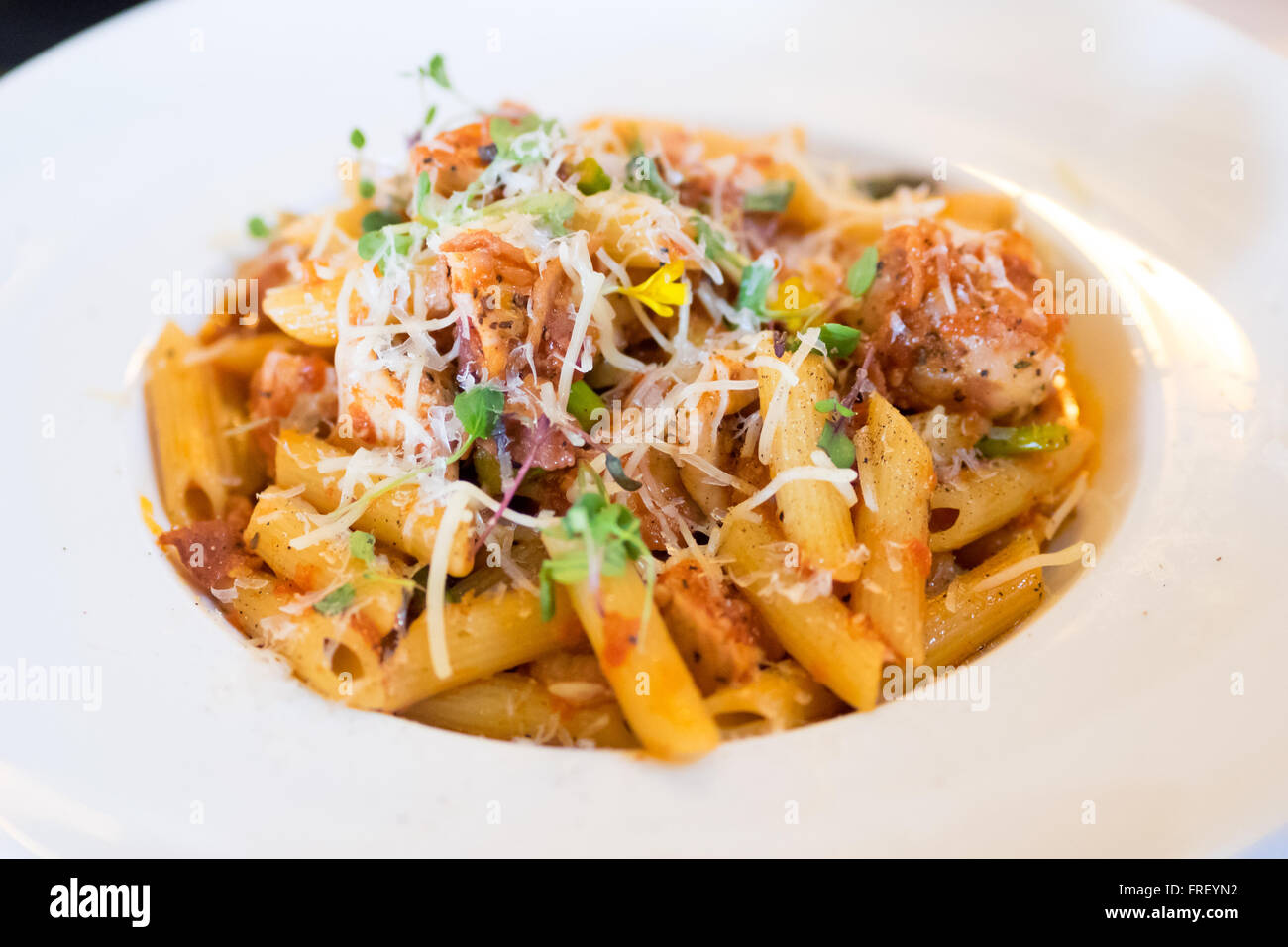 A plate of penne pollo (chicken) pasta in a tomato and red pepper sauce with Parmigiano Reggiano, boar bacon, and sprouts. Stock Photo