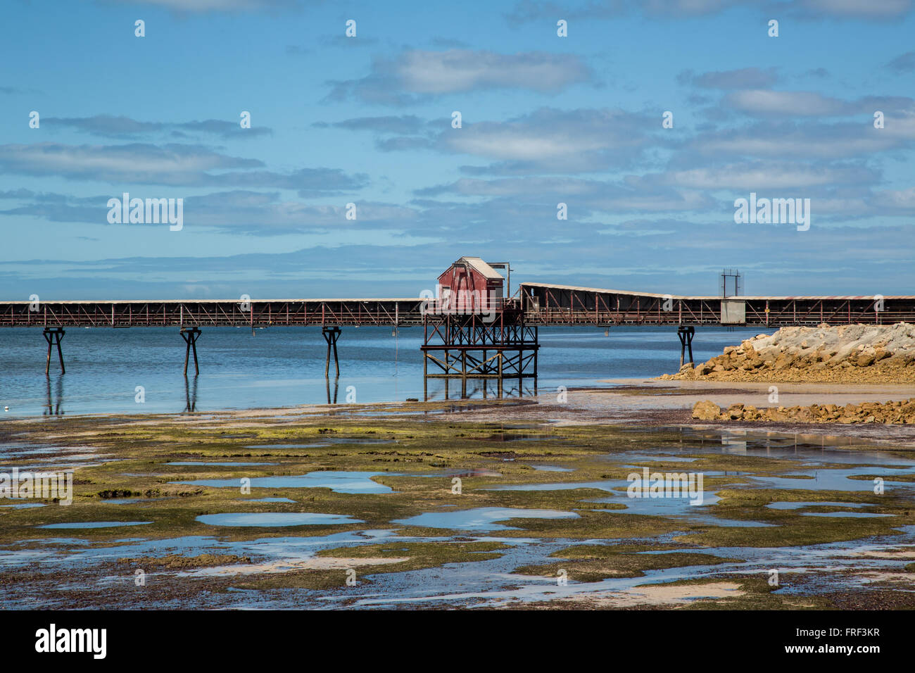 industrial loading jetty with red house, shallow water speckled with seaweed and sandbanks under blue sky with cumulus clouds Stock Photo