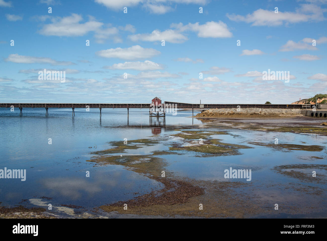 industrial loading jetty with red house, shallow water speckled with seaweed and sandbanks under blue sky with cumulus clouds Stock Photo