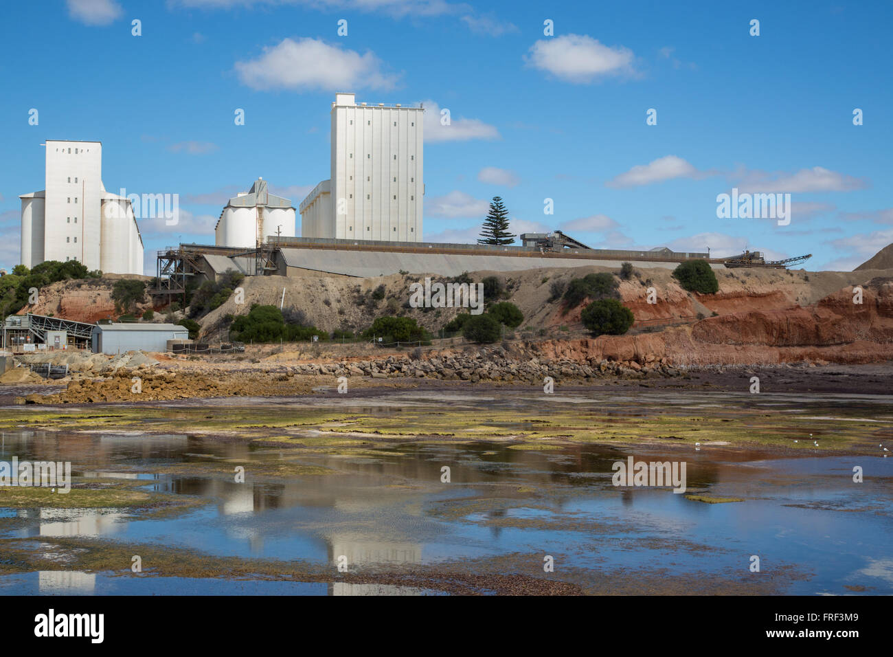 white crop storage silo facilities on top of sea shore with reflections in shallow water with mud and seaweed Stock Photo