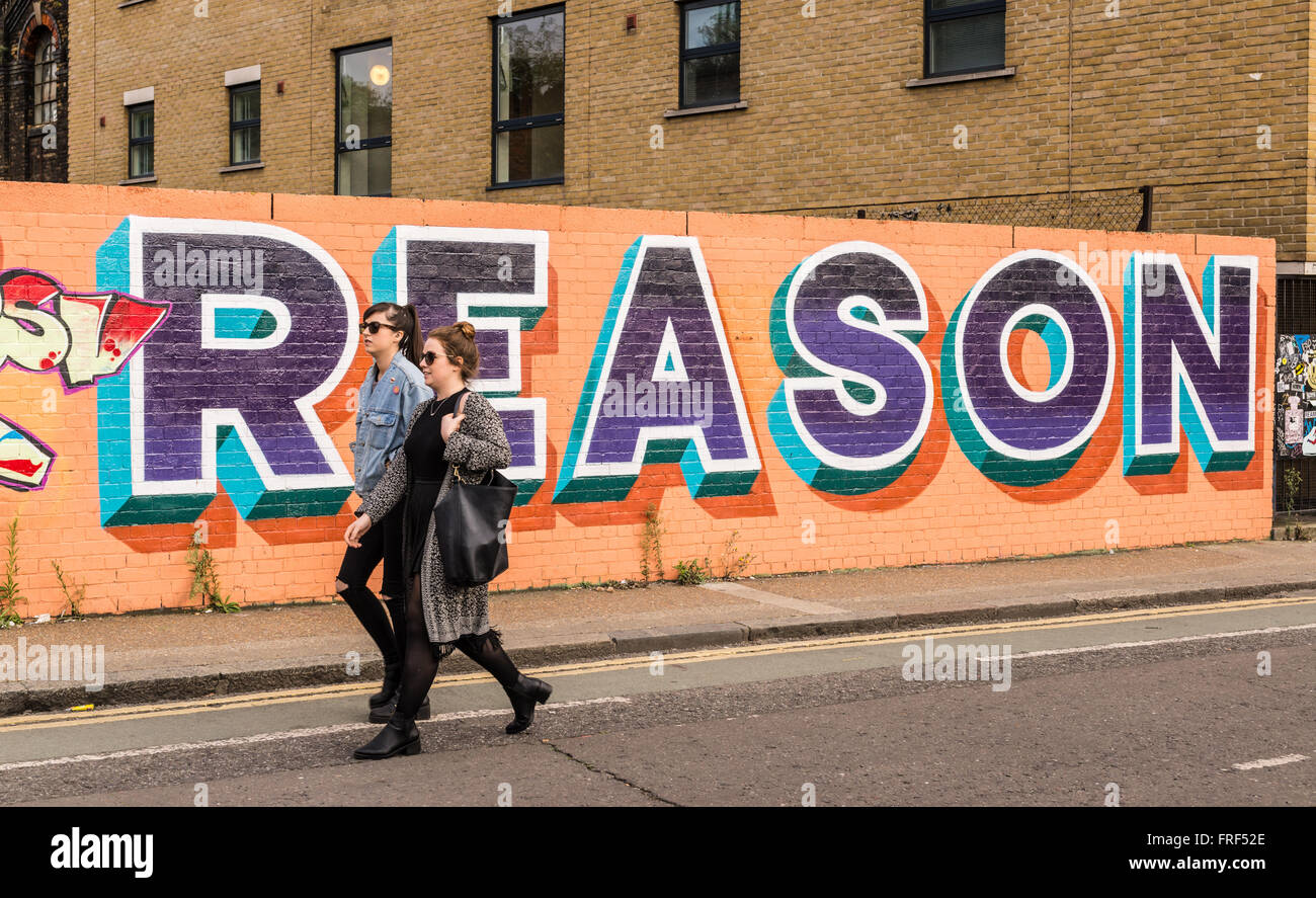 Two girls walking in front of a big graffiti with the word 'Reason' written on it. Stock Photo