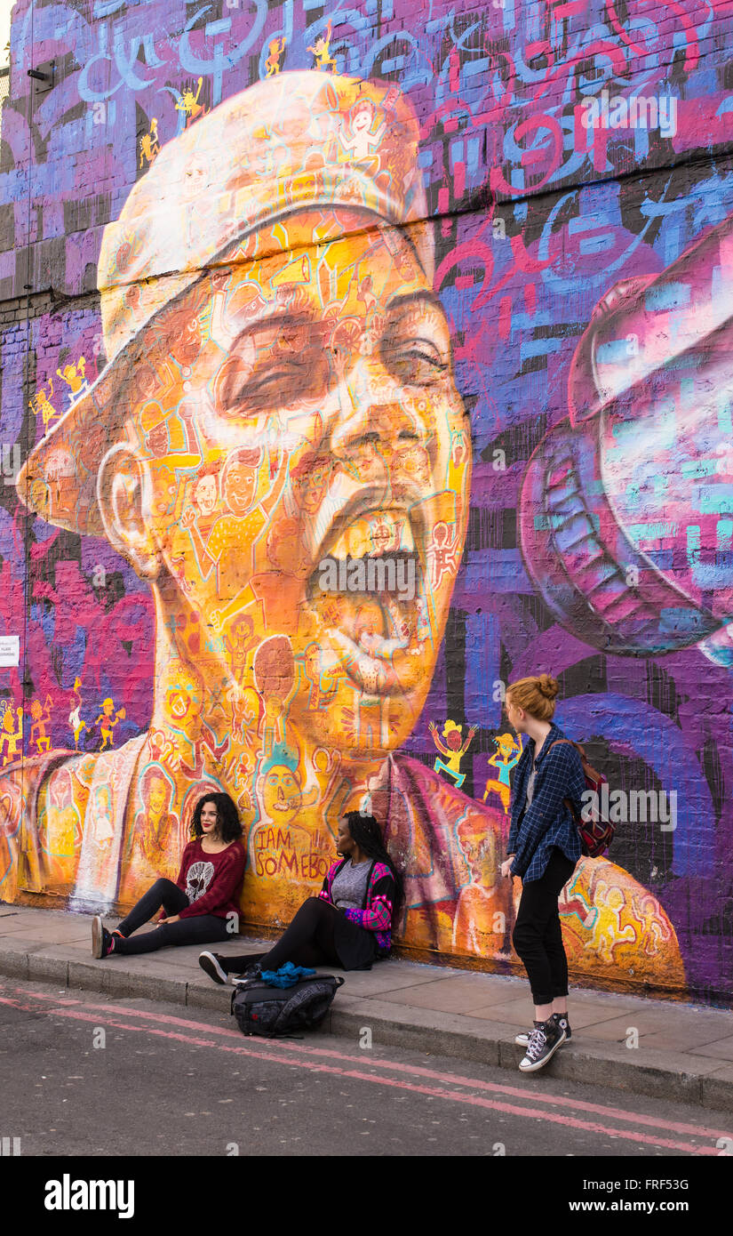 Three teenage girls standing in front of a wall with a big graffiti representing a boy shouting with a speaker. Stock Photo