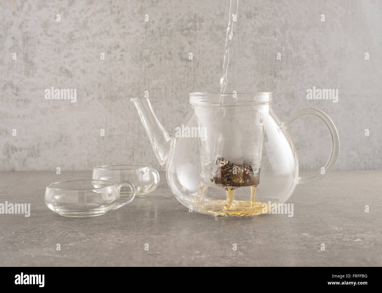 Hot water pouring in glass teapot and sifting through tea leaves. Two glass cups are placed on the side on a stone kitchen table Stock Photo