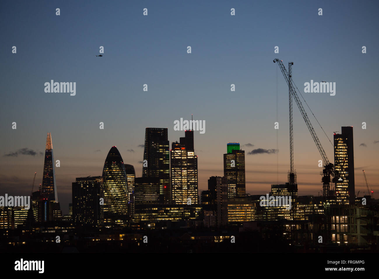 The sun is setting on the City of London and the lights are on, seen from Hackney, East London. A helicopter hovers above. Stock Photo