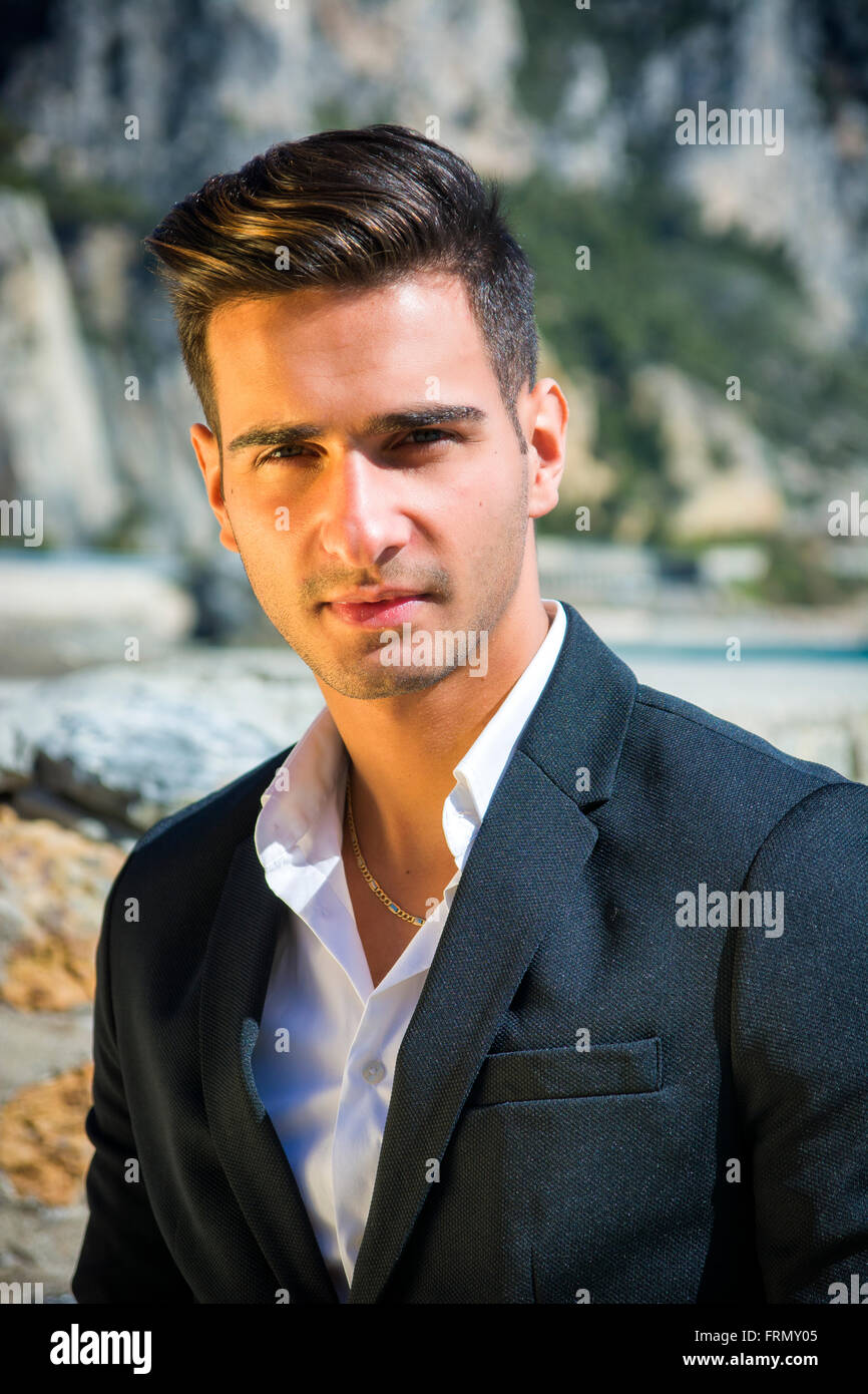 Young handsome man in classical suit on beach holding sunglasses while looking away. Sea waves on background Stock Photo