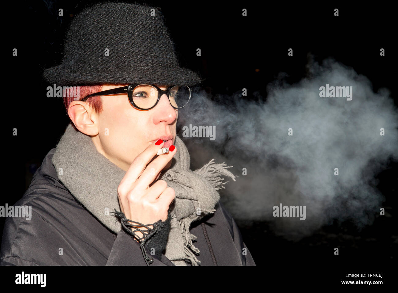 red-haired woman with hat and glasses standing outside and smoking a cigarette Stock Photo