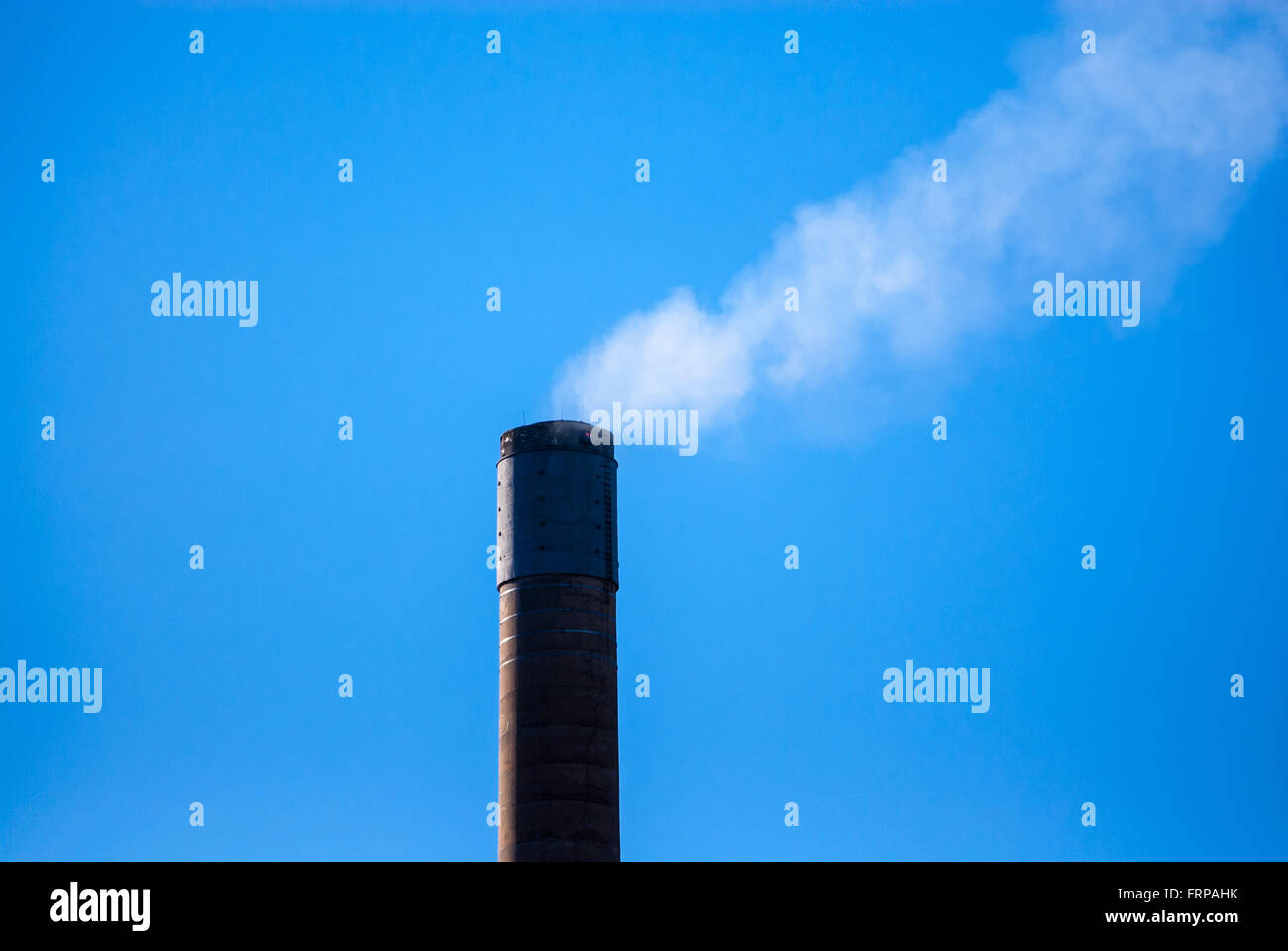 Top of dark industrial smoke stack releasing thin white smoke on clear blue sky. Stock Photo