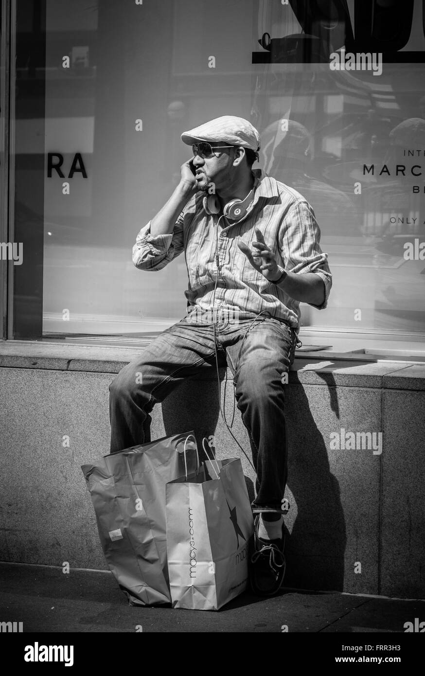 Young man sat outside shop talking on mobile phone with headphones and Macy's carrier bags, New York City, USA. Stock Photo