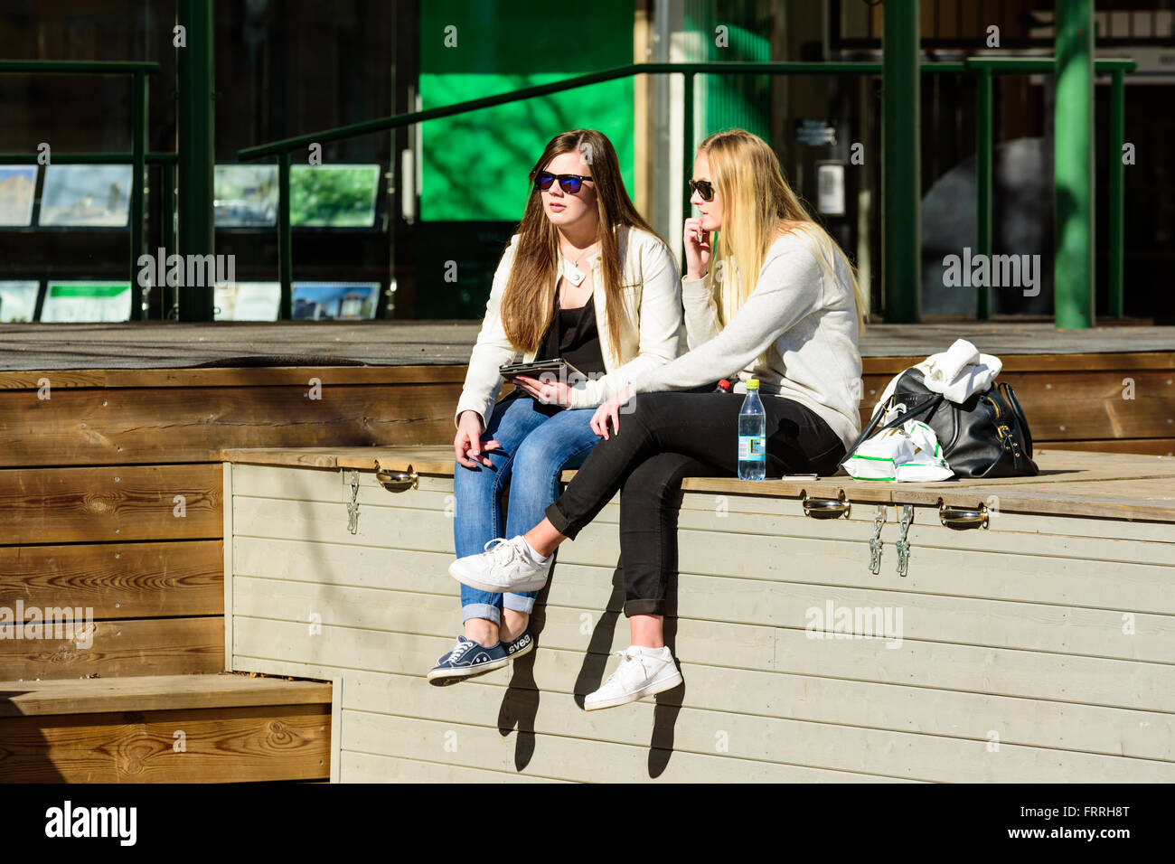 Kalmar, Sweden - March 17, 2016: Two female young adults sit on a wooden scene and are having a talk. Both wear sunglasses and b Stock Photo